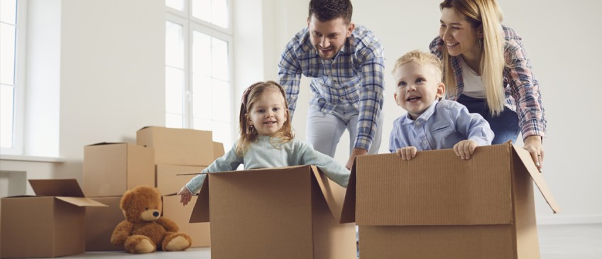 Ratgeber Miete oder Eigenheim - Eltern schieben Kinder in Umzugskartons durch Zimmer