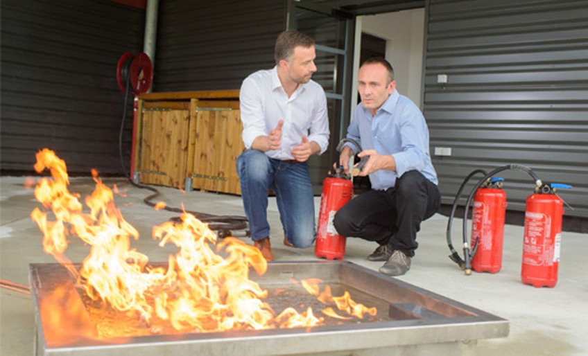 Verhalten im Brnadfall: Mann erklärt einem Anderen die Anwendung von Feuerlöschern.